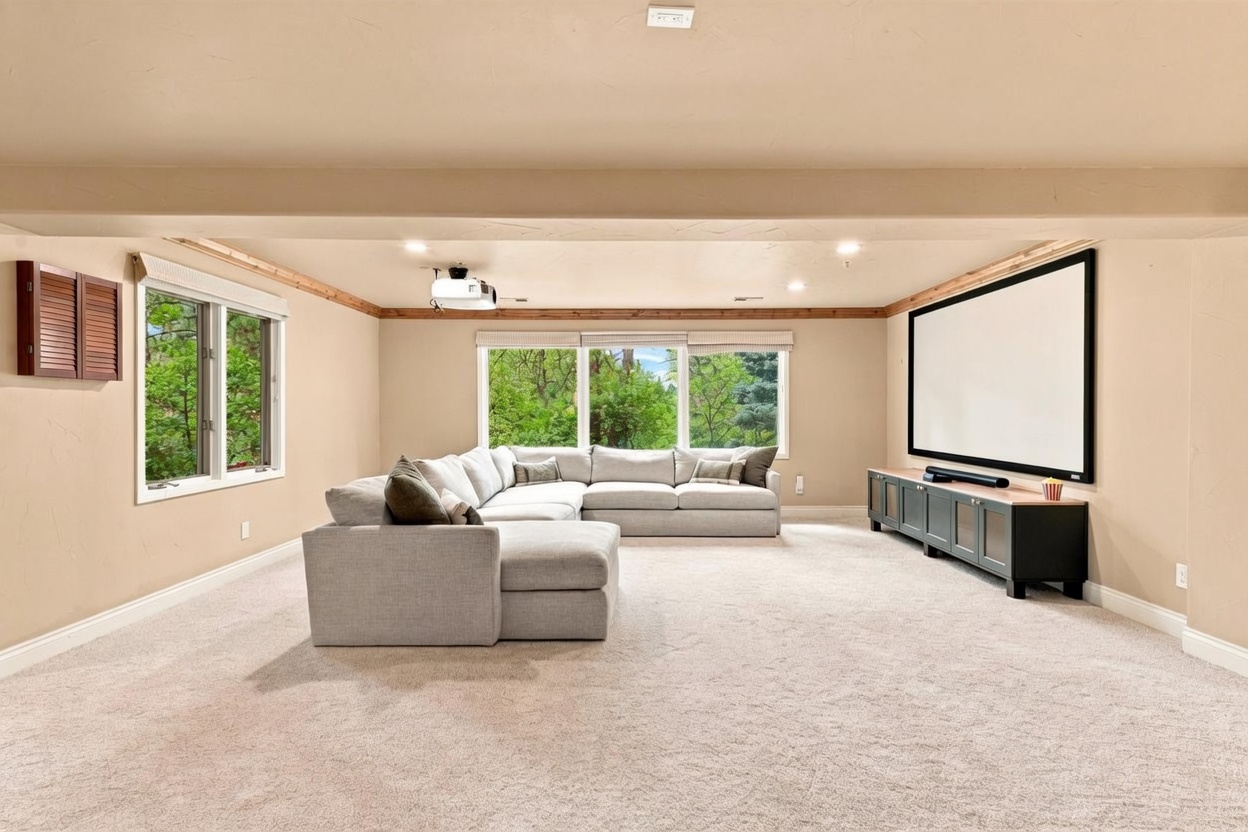 This spacious basement media room features a large, light-colored sectional sofa arranged for comfortable viewing of a projector screen mounted on the wall. The room is equipped with a ceiling-mounted projector and a dark media console, creating an ideal setup for entertainment. Large windows allow natural light to brighten the space, which is finished with neutral-toned walls and carpeting.