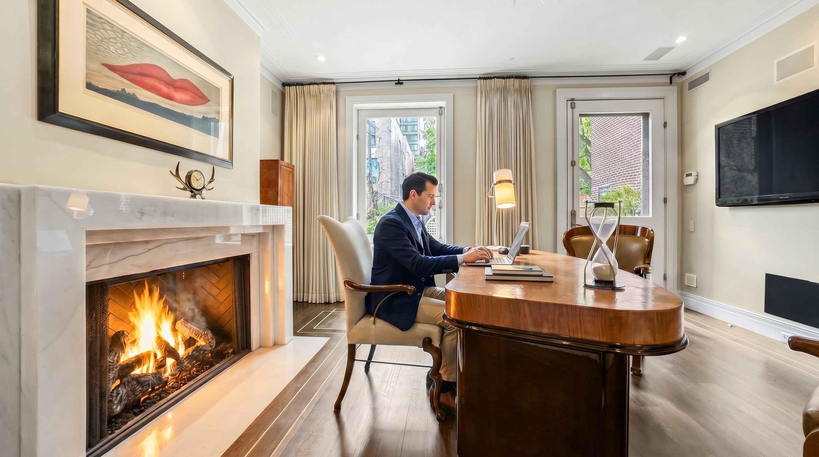 This sophisticated home office features a prominent desk with a curved top and a dark, polished base, positioned centrally in the room. To the left, a fireplace with a white stone surround and mantel serves as a focal point, topped by a large piece of art depicting lips. The room is illuminated by a star-shaped chandelier and natural light streaming through a window and a glass-paneled door that lead to an outdoor area.