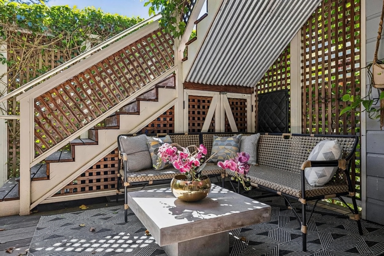 This cozy, covered outdoor seating area is tucked beneath a staircase, creating an intimate and shaded retreat. The space features a modern, low-profile sectional sofa with patterned throw pillows, a central concrete coffee table, and a geometric-patterned area rug. The backdrop is defined by decorative wooden lattice panels and a corrugated metal roof, offering a blend of rustic charm and contemporary design.