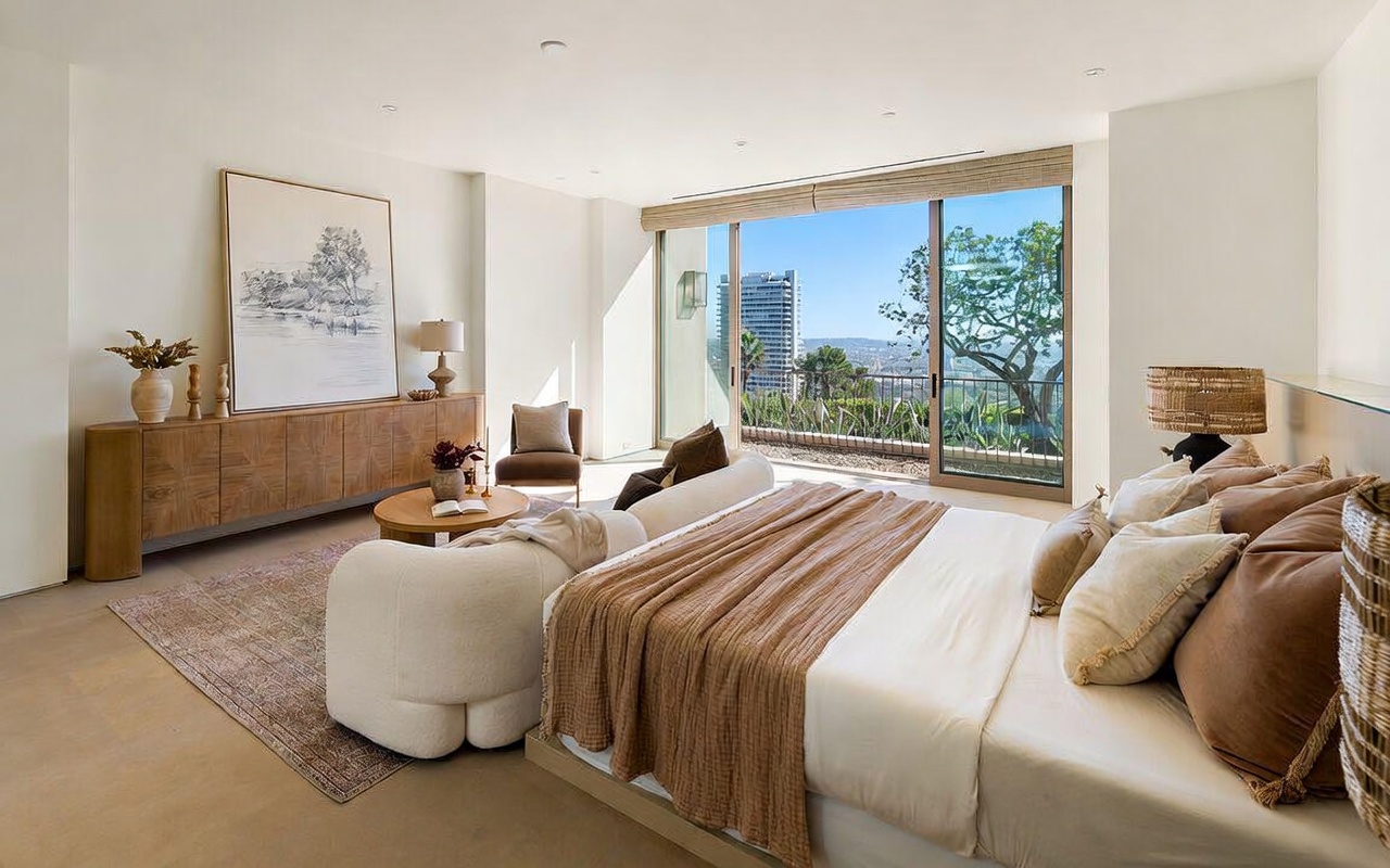 This luxurious primary bedroom features a sophisticated, neutral color palette with soft textures and a modern, minimalist aesthetic. A large bed with plush bedding sits prominently in the foreground, while a curved sofa and a wooden console table add depth to the seating area. The room is flooded with natural light from floor-to-ceiling sliding glass doors that open onto a balcony with expansive city and hillside views.