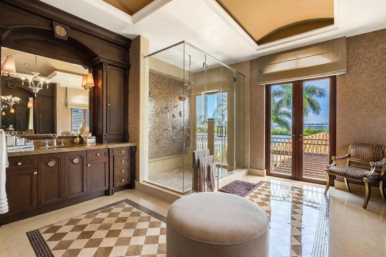 This luxurious primary bathroom features a sophisticated design with dark wood cabinetry, a large glass-enclosed walk-in shower with intricate mosaic tiling, and a central ottoman. The space is accented by elegant wallpaper, a double vanity with a large mirror, and French doors that open to a balcony, creating an airy and opulent atmosphere. The perspective is a wide-angle shot from the center of the room, emphasizing the high-end finishes and spacious layout.