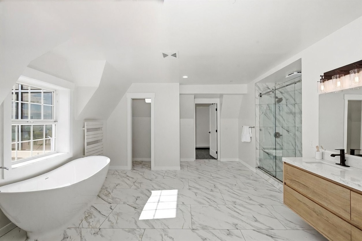 This bright and spacious primary bathroom features a freestanding soaking tub positioned near a large window and a glass-enclosed walk-in shower with patterned stone tile walls. The room is anchored by a light wood vanity with a modern faucet and a clean, minimalist aesthetic. The perspective captures the entire layout, highlighting the open floor plan and the transition between the bathing area and the adjacent rooms.