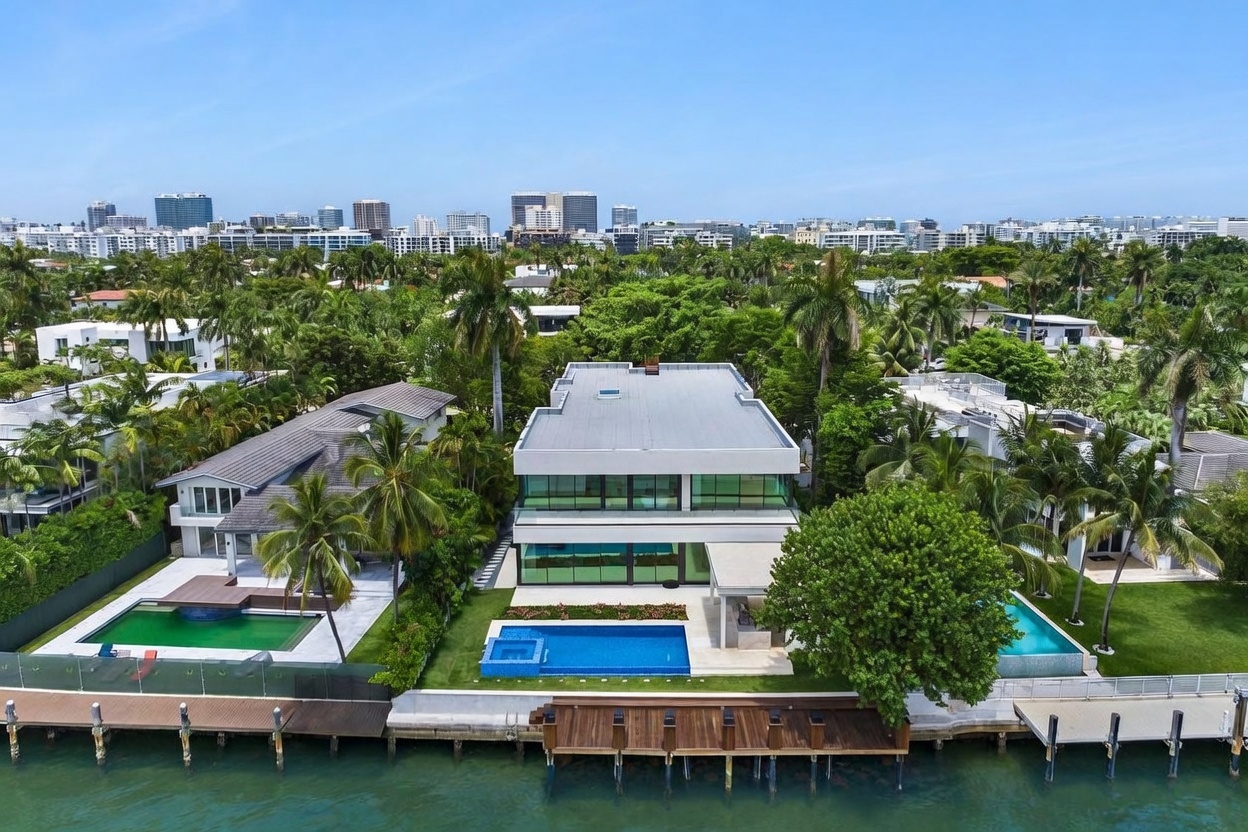 This high-angle aerial view captures a modern, multi-story waterfront residence featuring clean lines and extensive glass walls. The property includes a private swimming pool, a wooden dock extending into the water, and lush tropical landscaping. The perspective highlights the home's prime location along the canal, showcasing its contemporary architectural design and seamless integration with the outdoor living space.