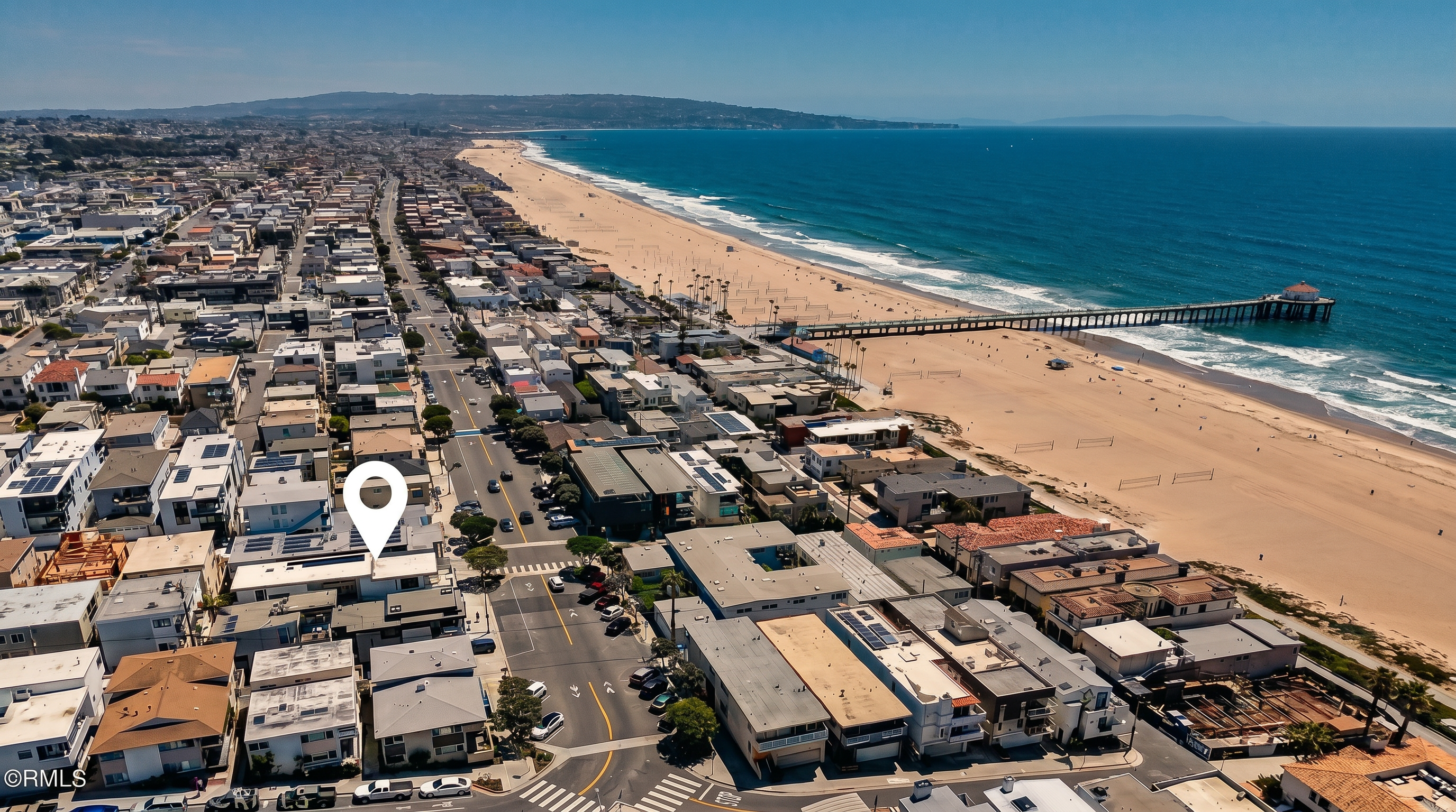 This high-angle aerial view captures a coastal neighborhood situated directly adjacent to a wide, sandy beach and the Pacific Ocean. A prominent red location pin highlights a specific property within the dense grid of residential homes, which are positioned just blocks from a long pier extending into the water. The perspective provides a clear sense of the property's prime location, emphasizing its proximity to the shoreline and the surrounding beach community.