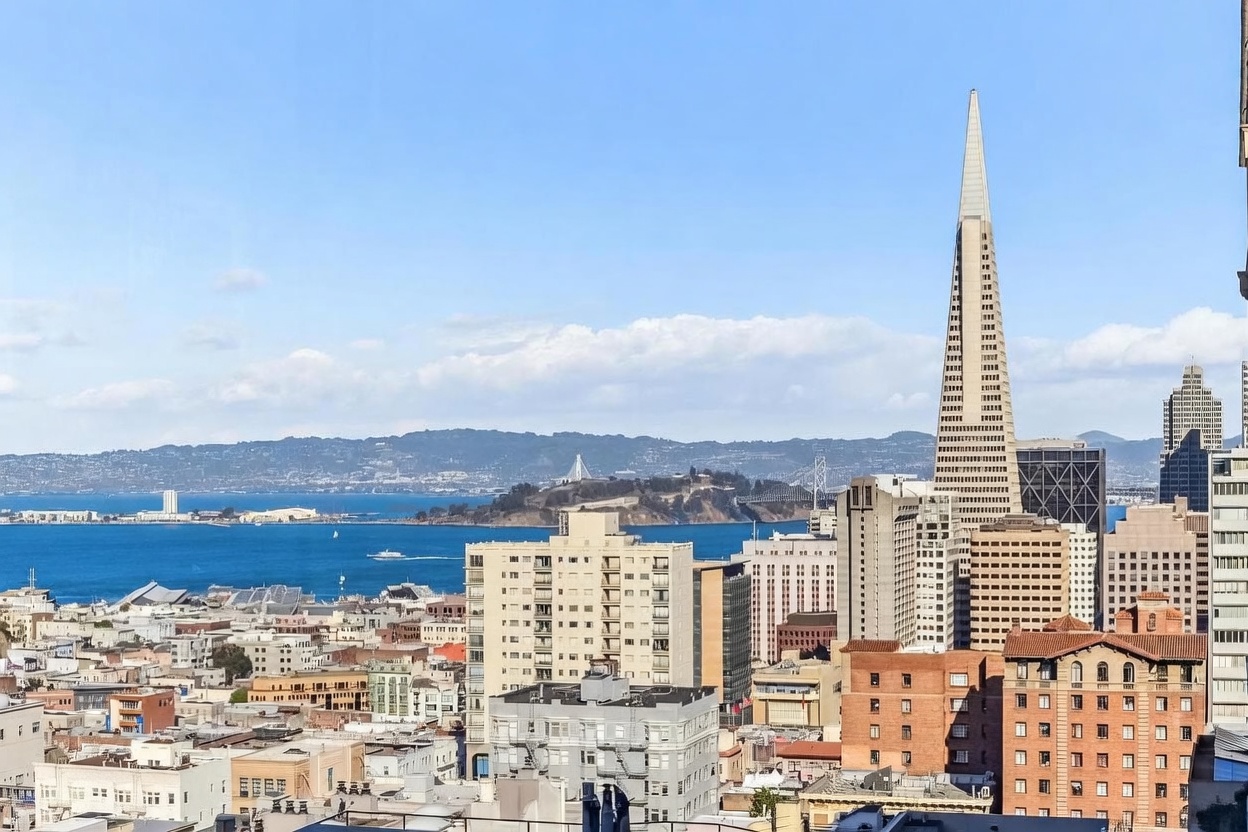 This high-angle aerial view captures a sweeping cityscape of San Francisco, prominently featuring the iconic Transamerica Pyramid rising above the dense urban skyline. The composition highlights the contrast between the historic and modern architecture of the city, with the sparkling blue waters of the bay and the distant hills providing a scenic backdrop. The perspective offers a clear sense of the property's prime location within the heart of the city's vibrant metropolitan landscape.