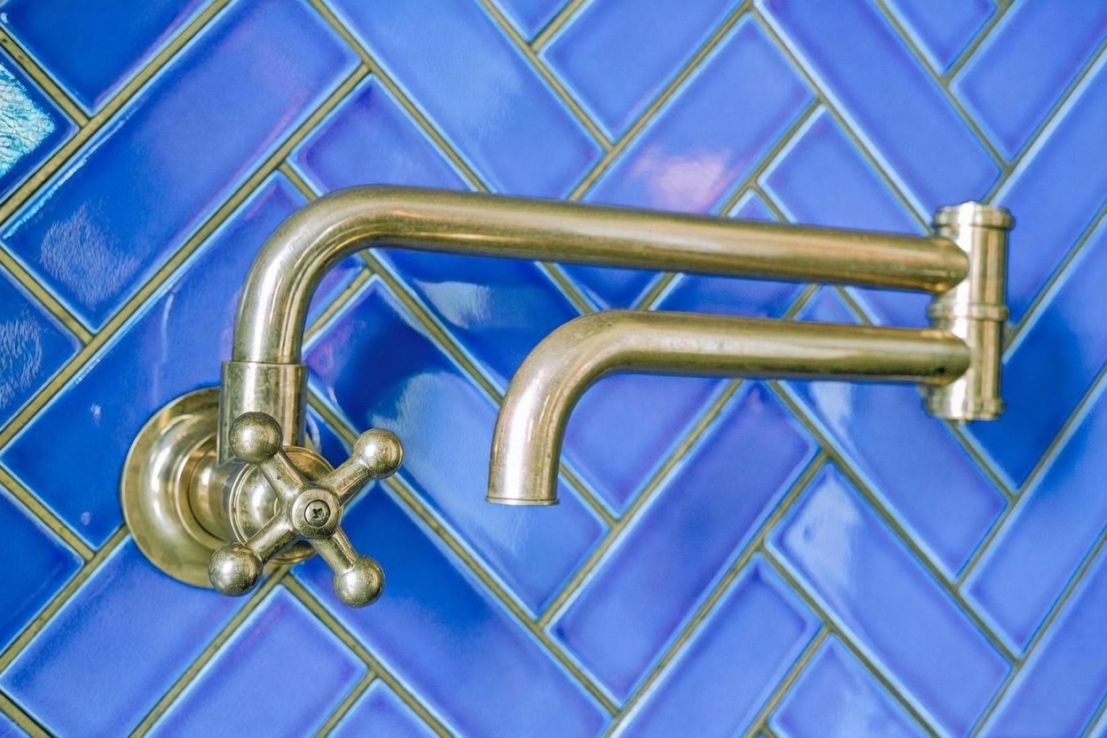 A close-up shot of a brass-toned, wall-mounted pot filler faucet featuring a classic cross-handle design. The fixture is set against a vibrant blue backsplash composed of rectangular tiles arranged in a herringbone pattern. The perspective is centered and direct, highlighting the metallic finish and the intricate tile work.
