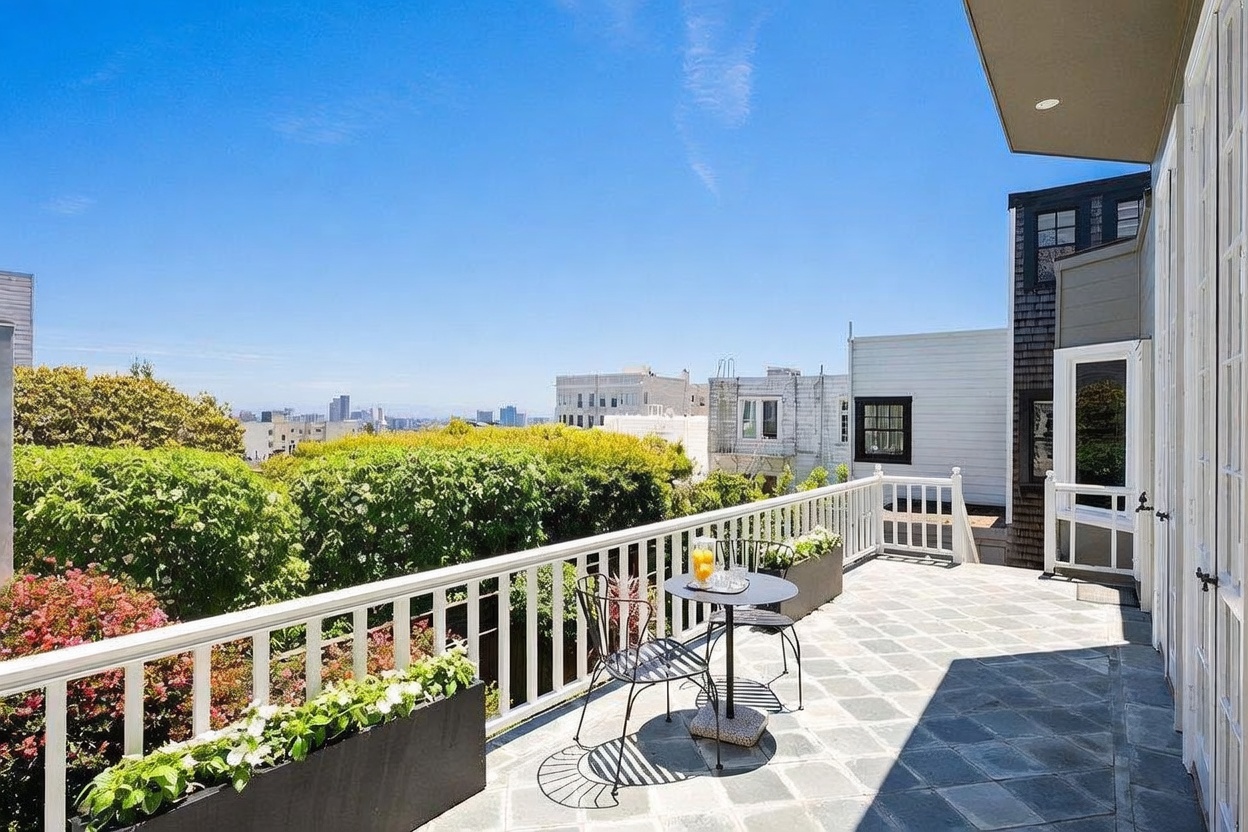 This elevated outdoor terrace features stone-tiled flooring and a white railing that overlooks a lush green hedge and a distant city skyline. A small bistro table with two chairs is positioned in the center, creating an inviting space for relaxation or morning coffee. The scene is captured from a slightly elevated perspective, emphasizing the open-air atmosphere and the scenic urban backdrop.