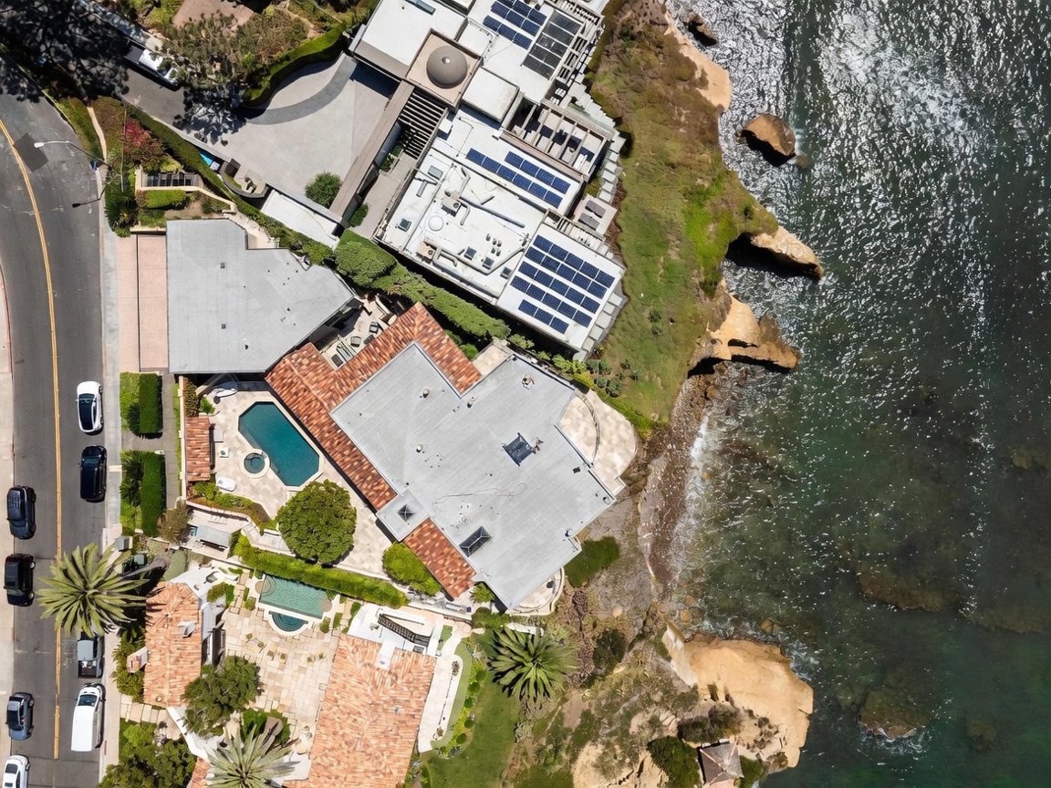 This high-angle aerial perspective captures a luxurious coastal property complex situated directly on a rocky cliffside overlooking the ocean. The estate features multiple residences with terracotta-tiled roofs, private swimming pools, and expansive stone-paved patios, all nestled against a rugged shoreline. A quiet residential street runs along the western edge, providing a clear view of the property's integration into the dramatic natural landscape.