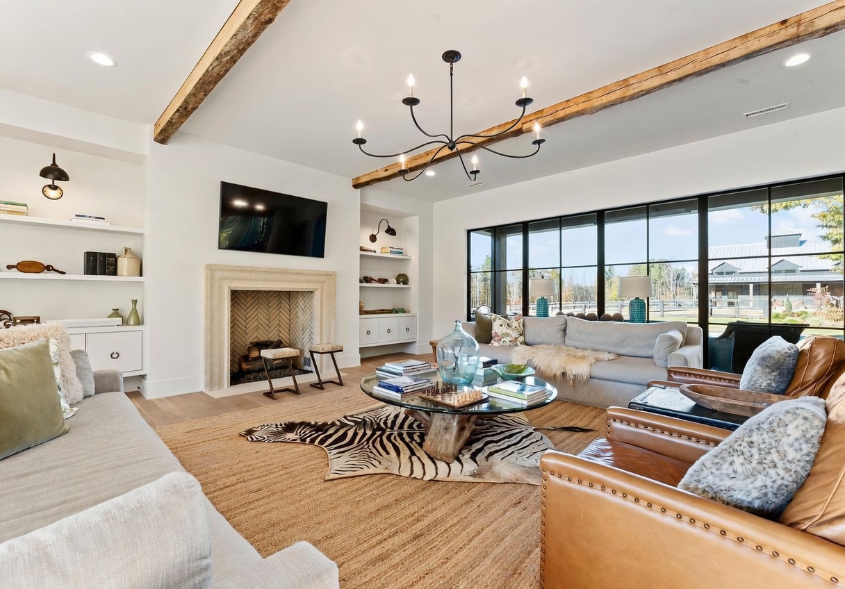 This spacious living room features a warm, inviting aesthetic with exposed wooden ceiling beams and a central stone fireplace surround. A large, circular glass coffee table sits atop a zebra-print rug, surrounded by a neutral-toned sofa and leather armchairs. Expansive black-framed windows provide a bright, airy feel and offer a view of the outdoor landscape.