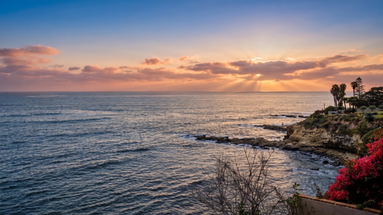 This view captures a stunning coastal panorama from an elevated vantage point, likely a balcony or terrace. The scene features a rugged rocky shoreline meeting the deep blue ocean under a bright, partly cloudy sky, with vibrant red bougainvillea flowers framing the foreground. The perspective offers a serene and expansive look at the natural landscape, highlighting the property's prime waterfront location.