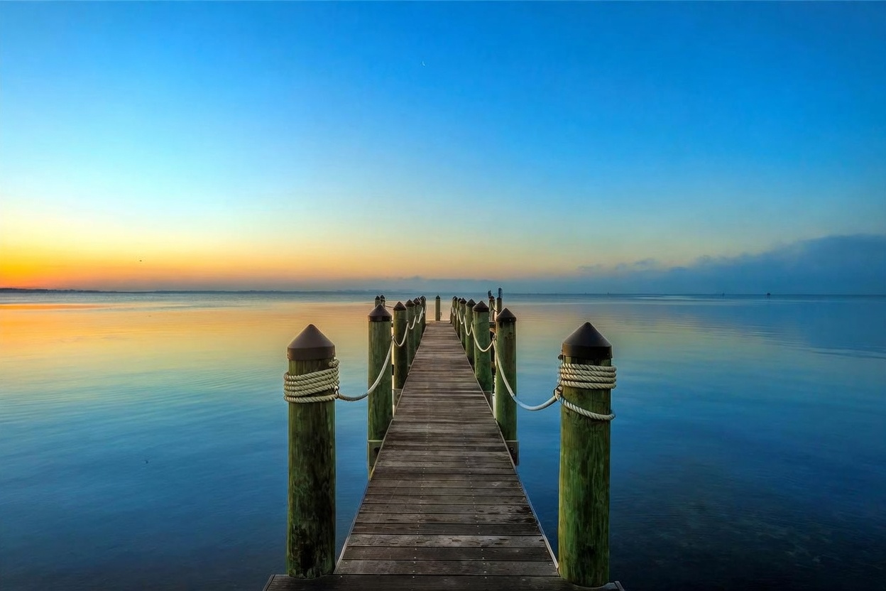 A serene, perspective-focused shot of a wooden pier extending into a calm, expansive body of water at sunrise. The scene features a symmetrical composition with weathered wooden pilings wrapped in nautical rope lining the walkway, leading the eye toward the horizon where the sky transitions from warm orange to cool blue. The water is mirror-like, reflecting the soft gradient of the morning light, creating a peaceful and tranquil atmosphere.