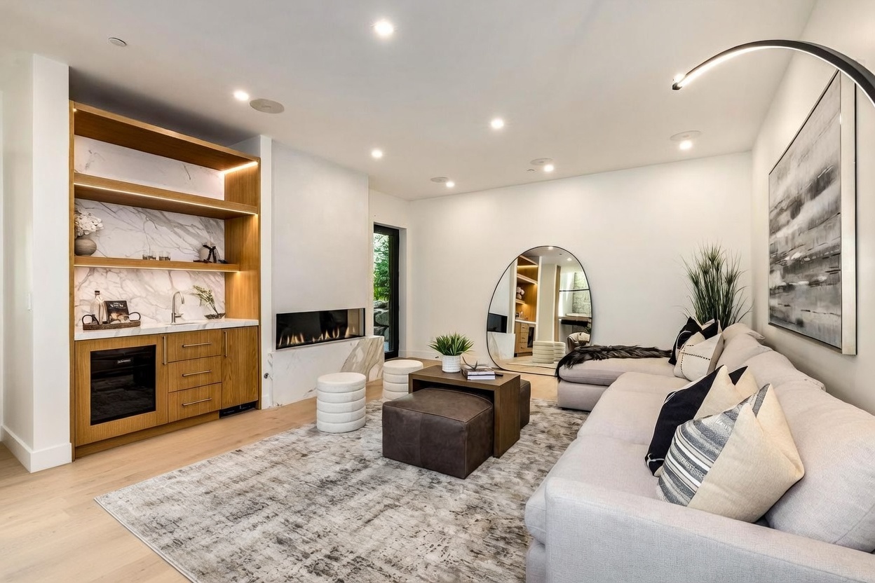This contemporary living room features a light-toned sectional sofa, two leather armchairs, and a central coffee table arrangement on a textured area rug. A built-in wet bar with light wood cabinetry and a stone-clad backsplash sits to the left, adjacent to a modern linear fireplace with a stone surround. The space is accented by a large, uniquely shaped floor mirror and a tall, arched floor lamp, creating a sophisticated and inviting atmosphere.