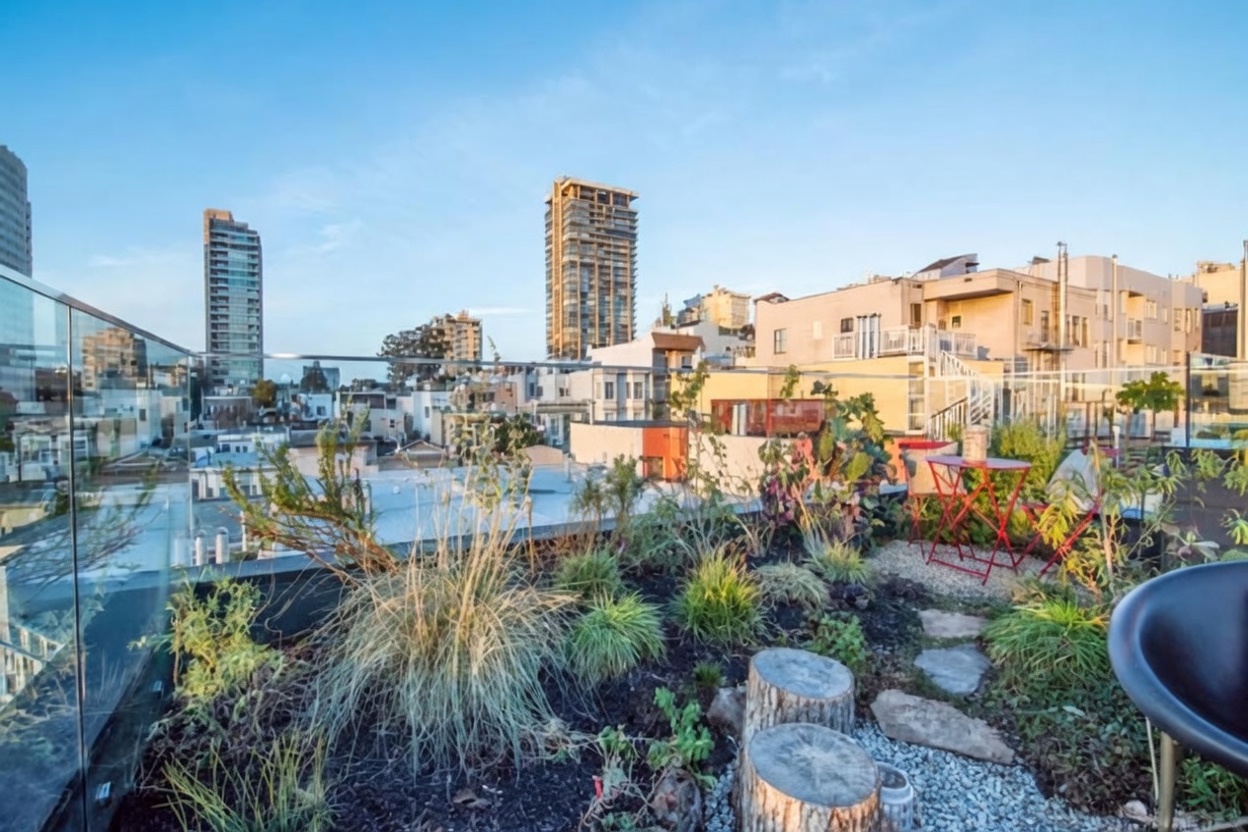 This rooftop terrace offers a panoramic urban view, featuring a landscaped garden area with native grasses, succulents, and stone accents. A glass railing provides an unobstructed perspective of the surrounding city skyline and neighboring buildings. A small bistro set with red chairs sits on the gravel surface, creating an inviting outdoor space for relaxation.