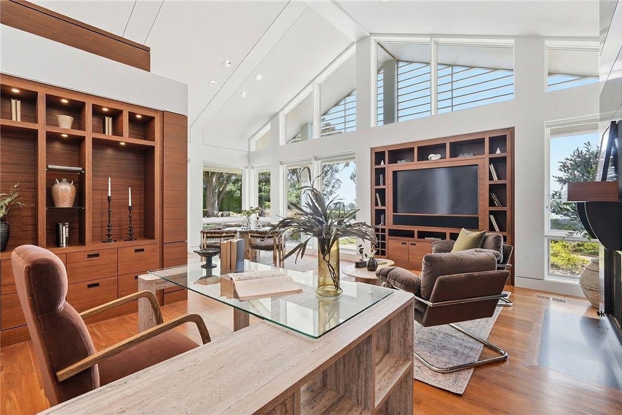 This sophisticated home office features a prominent glass-topped desk with a light wood base, positioned to overlook the living area. The room is defined by rich wood built-in cabinetry, a vaulted ceiling with large windows that flood the space with natural light, and a comfortable seating area centered around a television. The open-concept layout creates a seamless flow between the workspace and the lounge, offering a bright and airy atmosphere with views of the outdoors.