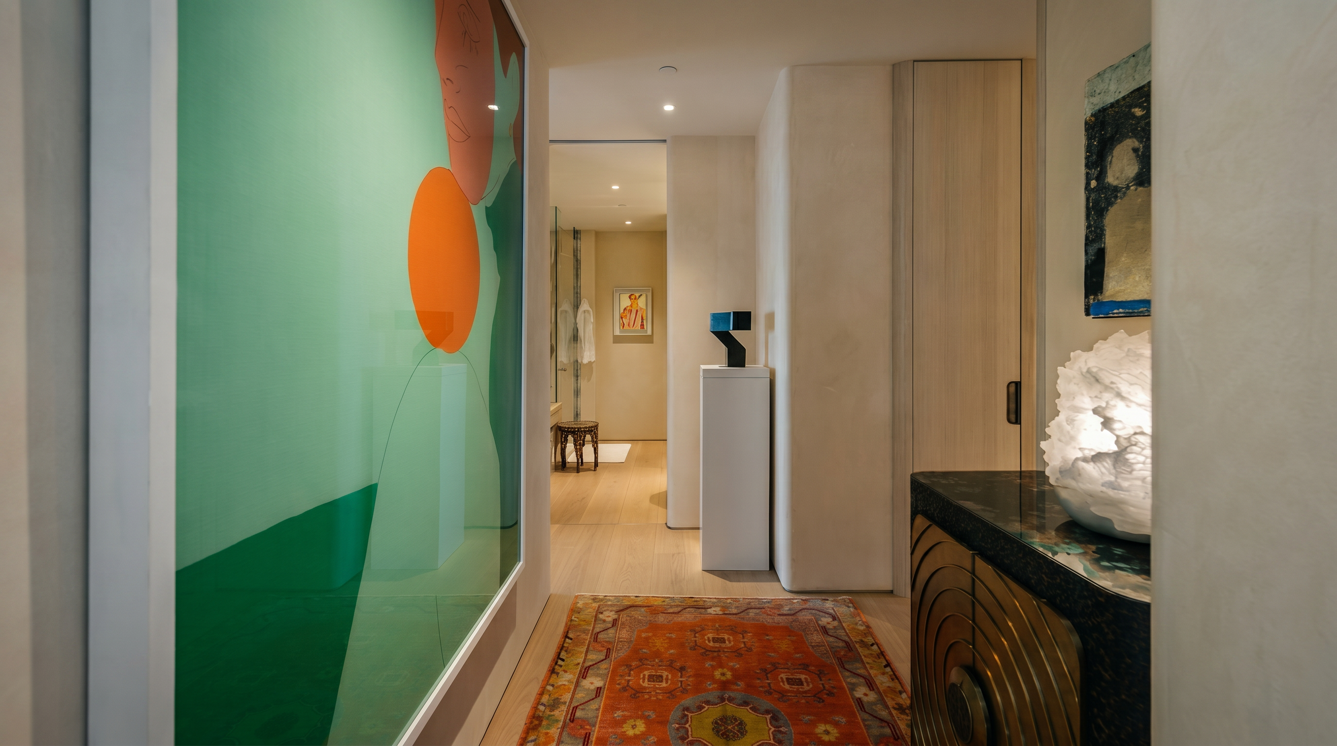 This elegant hallway features a vibrant, large-scale abstract art piece on the left wall, balanced by a warm, intricately patterned area rug that draws the eye toward the back of the home. To the right, a textured console table with a dark stone top and a sculptural lamp adds a sophisticated touch, while a minimalist pedestal display stands further down the corridor. The perspective looks through the hallway into a bright, neutral-toned space, creating a sense of depth and refined modern luxury.