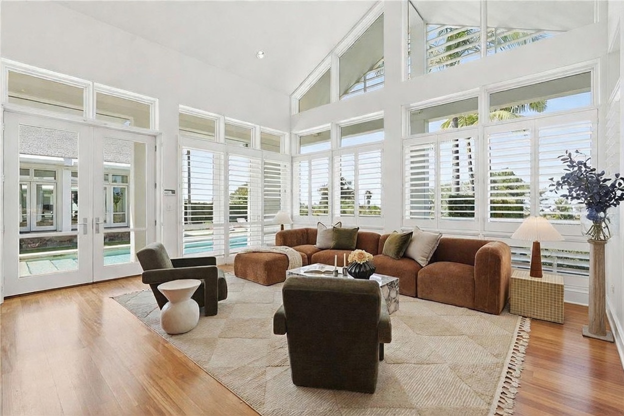 This bright and airy living room features a vaulted ceiling with expansive windows that allow for abundant natural light. The space is furnished with a large, curved brown sofa, a central coffee table, and two armchairs arranged on a textured area rug over light wood flooring. The room offers a seamless connection to the outdoors through glass doors, creating a sophisticated and inviting atmosphere.