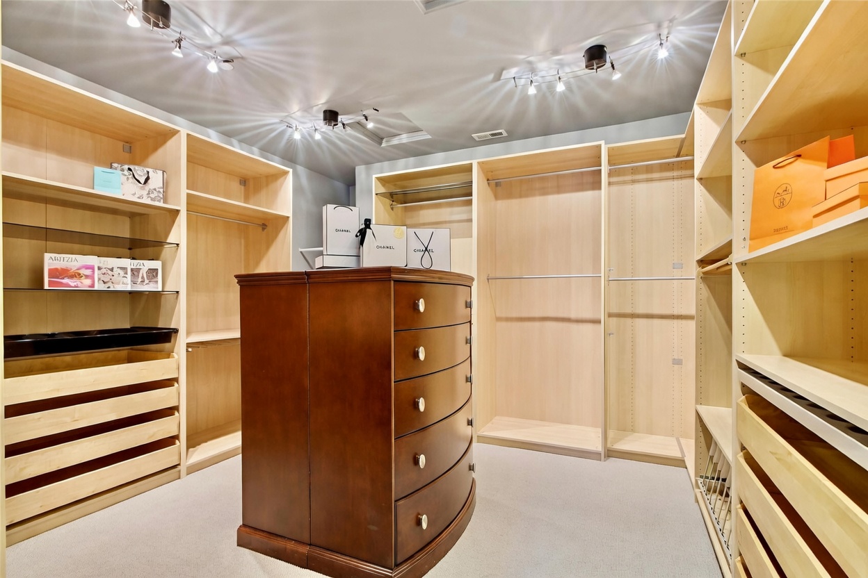 This spacious walk-in closet features extensive light wood shelving and hanging rods lining the walls, providing ample storage and organization space. A central dark wood dresser with multiple drawers serves as a focal point, while recessed track lighting illuminates the room from above. The neutral carpet and clean, organized layout create a sophisticated and functional dressing area.