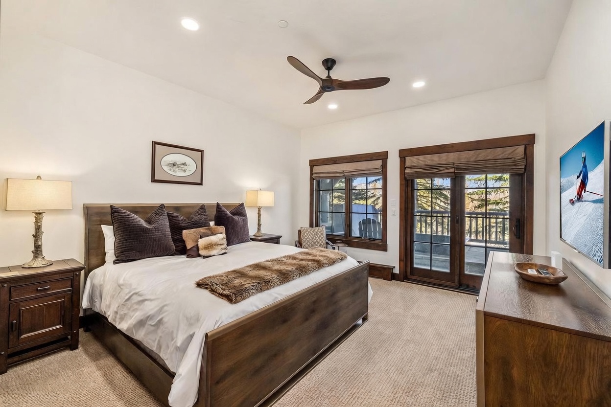 This primary bedroom features a warm, rustic aesthetic with a large wooden bed frame topped with dark pillows and a faux-fur throw. The room is illuminated by bedside lamps and recessed lighting, while a television mounted on the wall displays a mountain scene. Large glass doors provide access to a private balcony, allowing natural light to fill the space and offering a view of the outdoors.