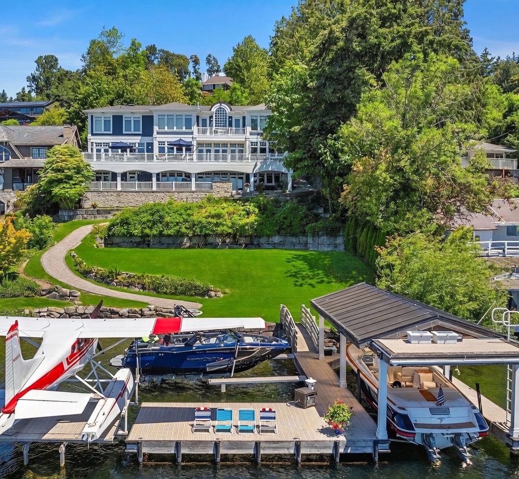 This stunning aerial view captures a luxurious waterfront estate featuring a multi-level residence with expansive decks overlooking a meticulously manicured lawn. The property includes a private dock equipped with a seaplane, a sleek motorboat, and a covered boat slip, highlighting an exclusive lifestyle. The lush greenery and serene water setting create an inviting and high-end atmosphere perfect for waterfront living.