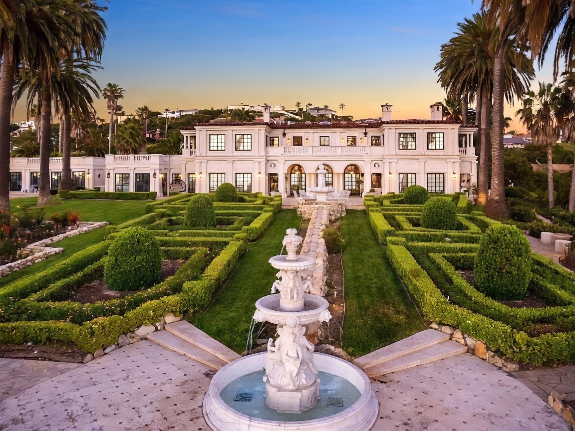 This grand estate features a symmetrical, classical architectural design with a light-colored facade and a multi-level structure. A formal, manicured garden with intricate boxwood hedges and a central tiered stone fountain serves as the focal point of the expansive backyard. The scene is captured from an elevated perspective, showcasing the impressive scale of the residence against a clear sky at dusk.