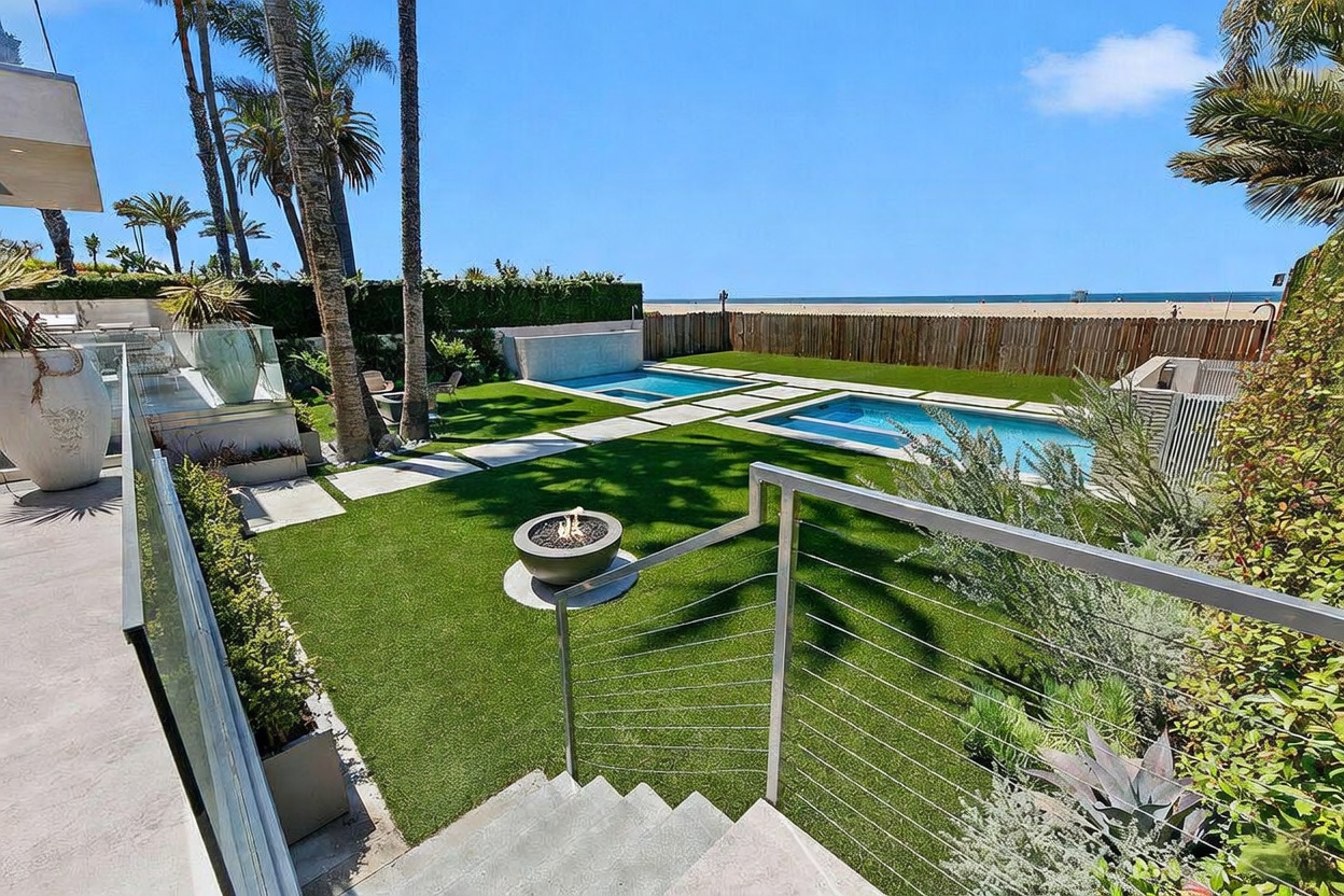 This elevated perspective captures a luxurious backyard oasis featuring a manicured lawn, a rectangular swimming pool, and a separate spa. A modern fire pit sits prominently in the foreground, while a sleek metal railing overlooks the space, which transitions seamlessly toward a beach and ocean view in the distance. The scene is framed by tall palm trees and lush landscaping, creating a serene and high-end coastal atmosphere.