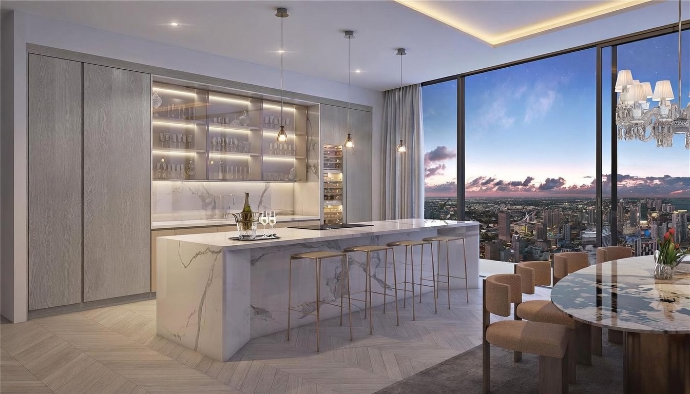 This modern, high-end kitchen features a large stone-topped island with bar seating and integrated cabinetry with illuminated glass shelving. The space is framed by expansive floor-to-ceiling windows that offer a panoramic view of the city skyline at dusk. A dining area with a circular table is visible in the foreground, contributing to an open-concept layout that emphasizes luxury and urban living.