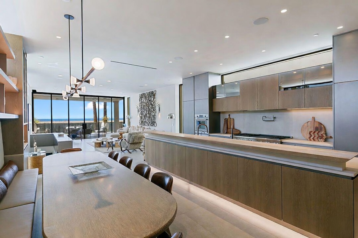 This open-concept space features a sleek, modern kitchen with light wood cabinetry and a long island that transitions into a dining area. Large glass doors at the far end provide an expansive view of the ocean, bathing the room in natural light. The minimalist aesthetic is complemented by a contemporary chandelier and recessed lighting, creating a sophisticated and airy atmosphere.