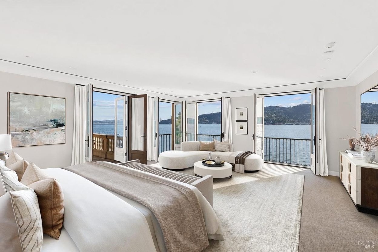 This spacious primary bedroom features a neutral color palette and a curved sofa arrangement positioned to take advantage of the panoramic water views. Large glass doors open onto a private balcony, flooding the room with natural light and creating a seamless indoor-outdoor connection. The room is furnished with a plush bed, a sleek dresser, and minimalist decor, offering a serene and luxurious atmosphere.