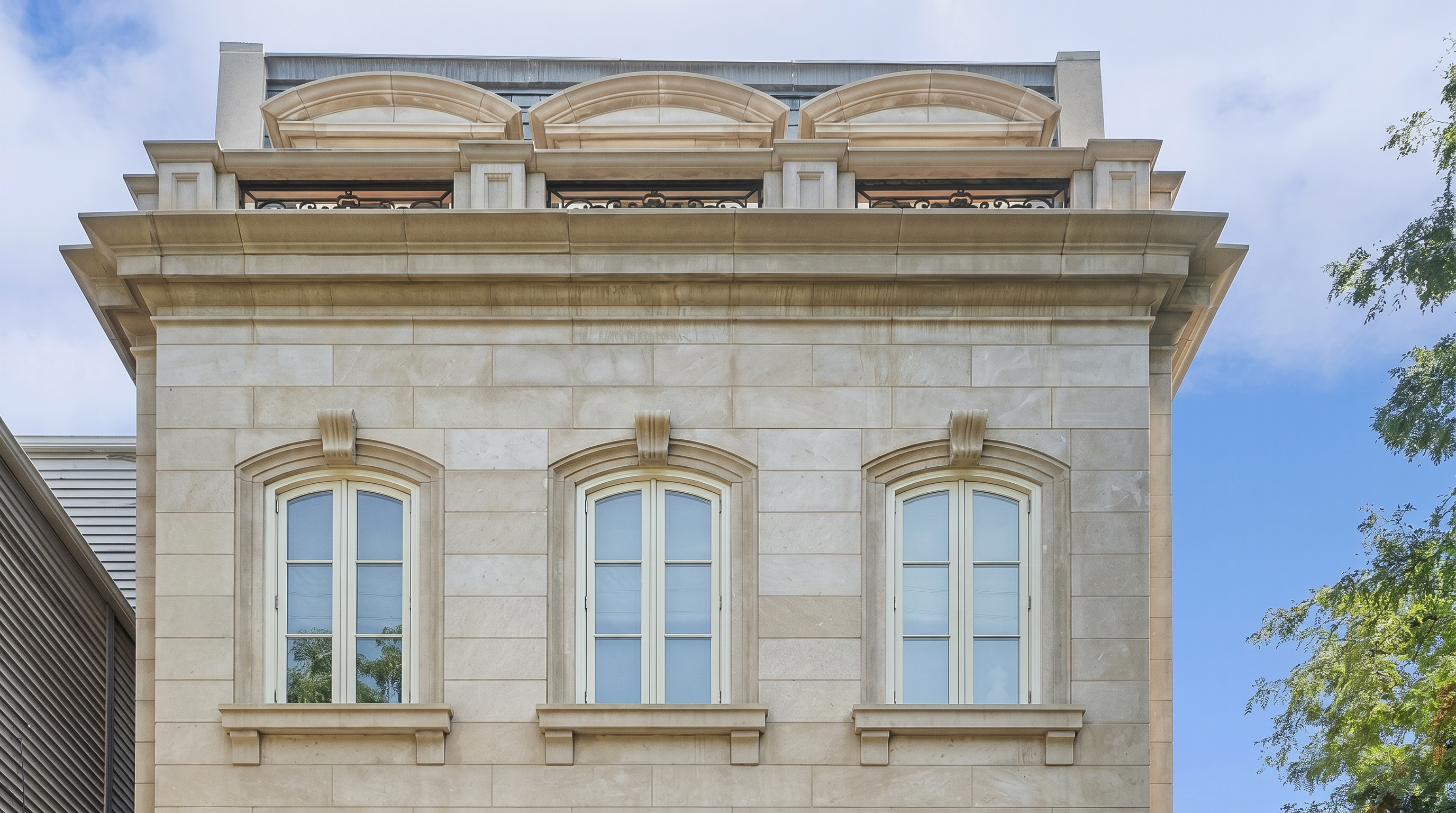 This image shows a stately stone facade of a multi-story residence, characterized by its classical architectural elements. It features three symmetrical arched windows on the upper level, decorative stone keystones, and a distinguished roofline with arched dormers. The facade's clean masonry work and refined proportions suggest a high-end luxury property.