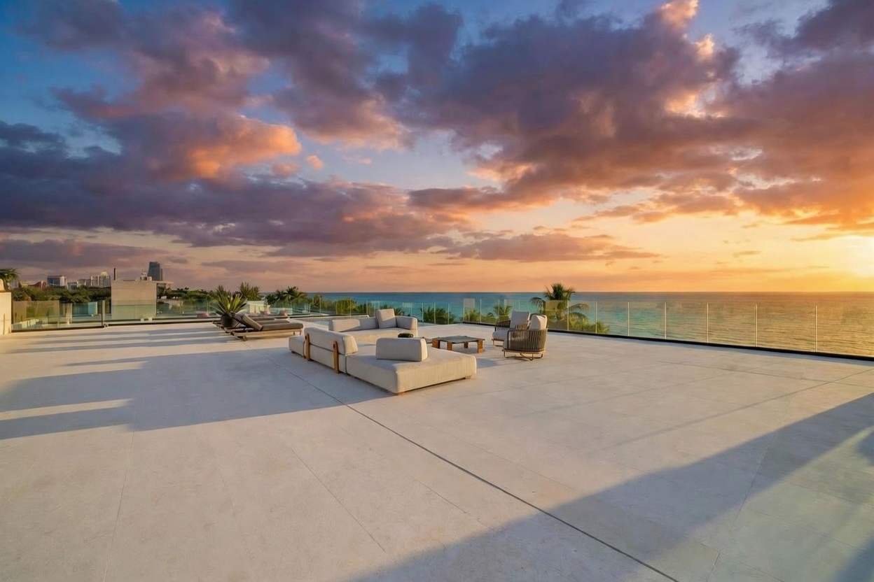 This expansive rooftop terrace offers a luxurious outdoor living space with panoramic ocean views at sunset. The area is furnished with contemporary modular seating and lounge chairs, set against a backdrop of a vibrant, cloud-filled sky and the horizon line. A sleek glass railing provides an unobstructed view, emphasizing the modern and sophisticated atmosphere of this high-end property.