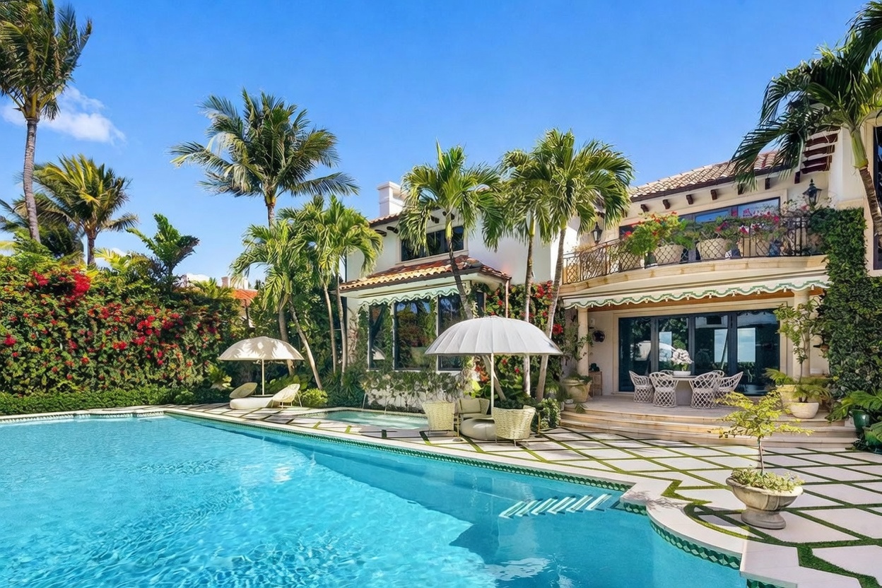 This luxurious backyard features a large, sparkling swimming pool as the centerpiece, surrounded by a geometric stone patio with integrated greenery. A covered patio area provides an elegant outdoor dining space, while a second-story balcony with wrought-iron railings overlooks the pool. The scene is framed by lush tropical landscaping, including tall palm trees and vibrant flowering bushes, creating a serene and private resort-like atmosphere.
