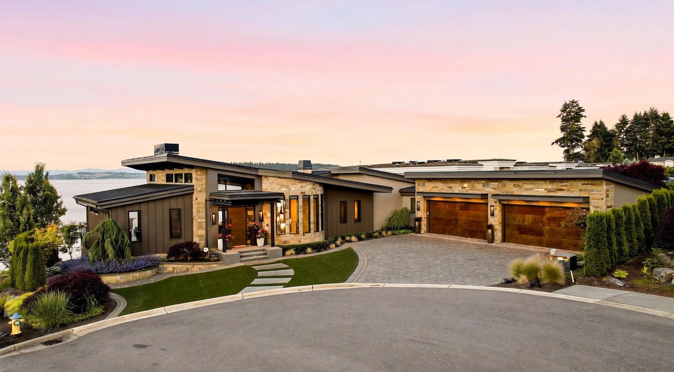 This contemporary luxury residence features a striking architectural design with a mix of dark metal siding and textured stone accents. The front entryway is highlighted by a warm wood door and a covered porch, while a paved driveway leads to a spacious multi-car garage. Set against a sunset sky, the home overlooks a body of water, with manicured landscaping and a curved stone walkway completing the elegant curb appeal.