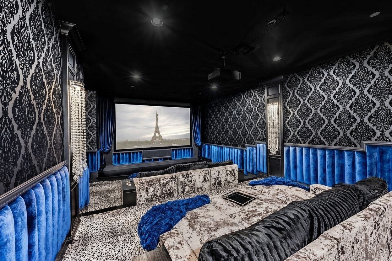 This opulent home theater features a dramatic, high-contrast design with deep blue velvet wall paneling and intricate black-and-white damask wallpaper. The room is furnished with plush, patterned seating and accented by sparkling crystal wall sconces that provide a luxurious, cinematic atmosphere. A large projection screen displays a scenic view of the Eiffel Tower, anchoring the space as a sophisticated entertainment retreat.