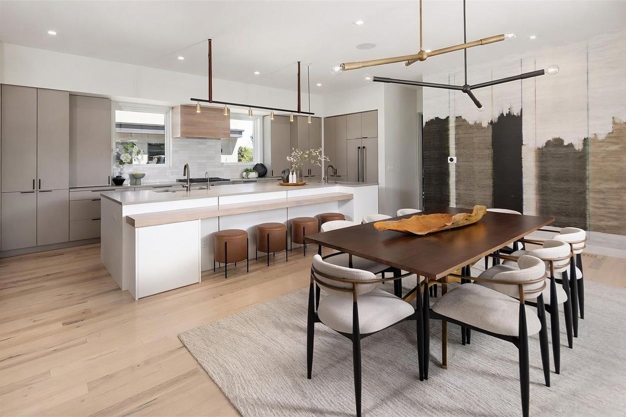 This open-concept kitchen and dining area features a large central island with a light wood breakfast bar and matching stools, complemented by sleek, neutral-toned cabinetry. A dark wood dining table sits on a textured area rug, illuminated by a modern, geometric chandelier, while a large-scale abstract mural adds a dramatic artistic focal point to the wall. The space is bright and airy, characterized by light wood flooring and a clean, contemporary aesthetic.