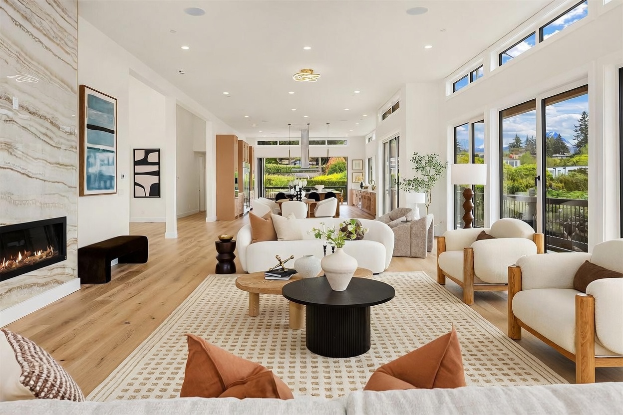 This expansive, open-concept living area features a light-toned wood floor and a prominent stone-clad chimney breast housing a linear fireplace. The space is furnished with a curved sofa and matching armchairs arranged on a textured area rug, creating a sophisticated and airy atmosphere. Large windows and sliding glass doors provide abundant natural light and a seamless transition to the outdoor deck, while the view extends toward the dining area and kitchen in the background.