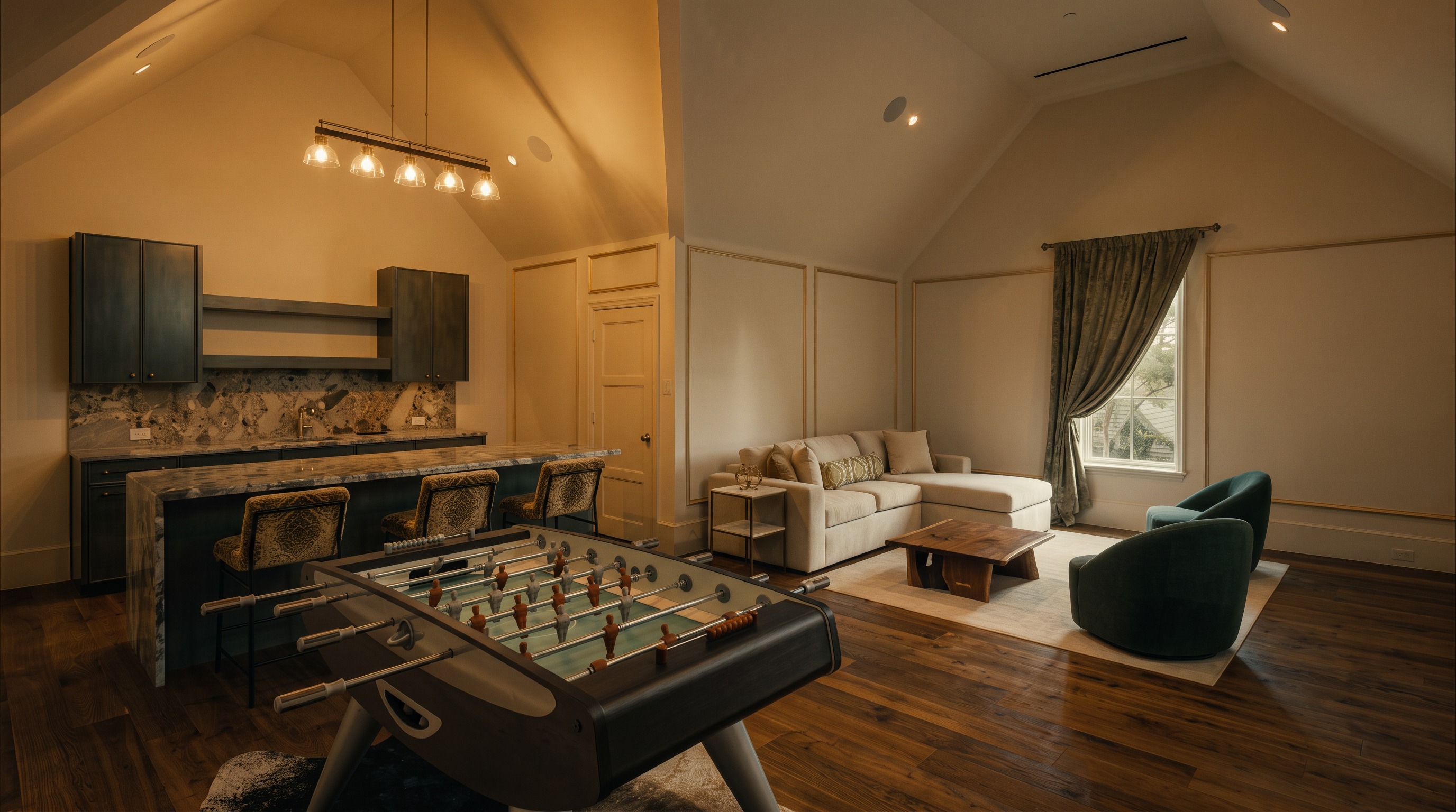 This spacious, open-concept living area features a vaulted ceiling and a dedicated game zone with a foosball table in the foreground. The room includes a wet bar with dark cabinetry and stone countertops, alongside a comfortable seating area with a light-colored sectional and two dark green armchairs. The space is finished with warm-toned wood flooring and neutral wall paneling, creating a sophisticated yet inviting atmosphere for entertaining.