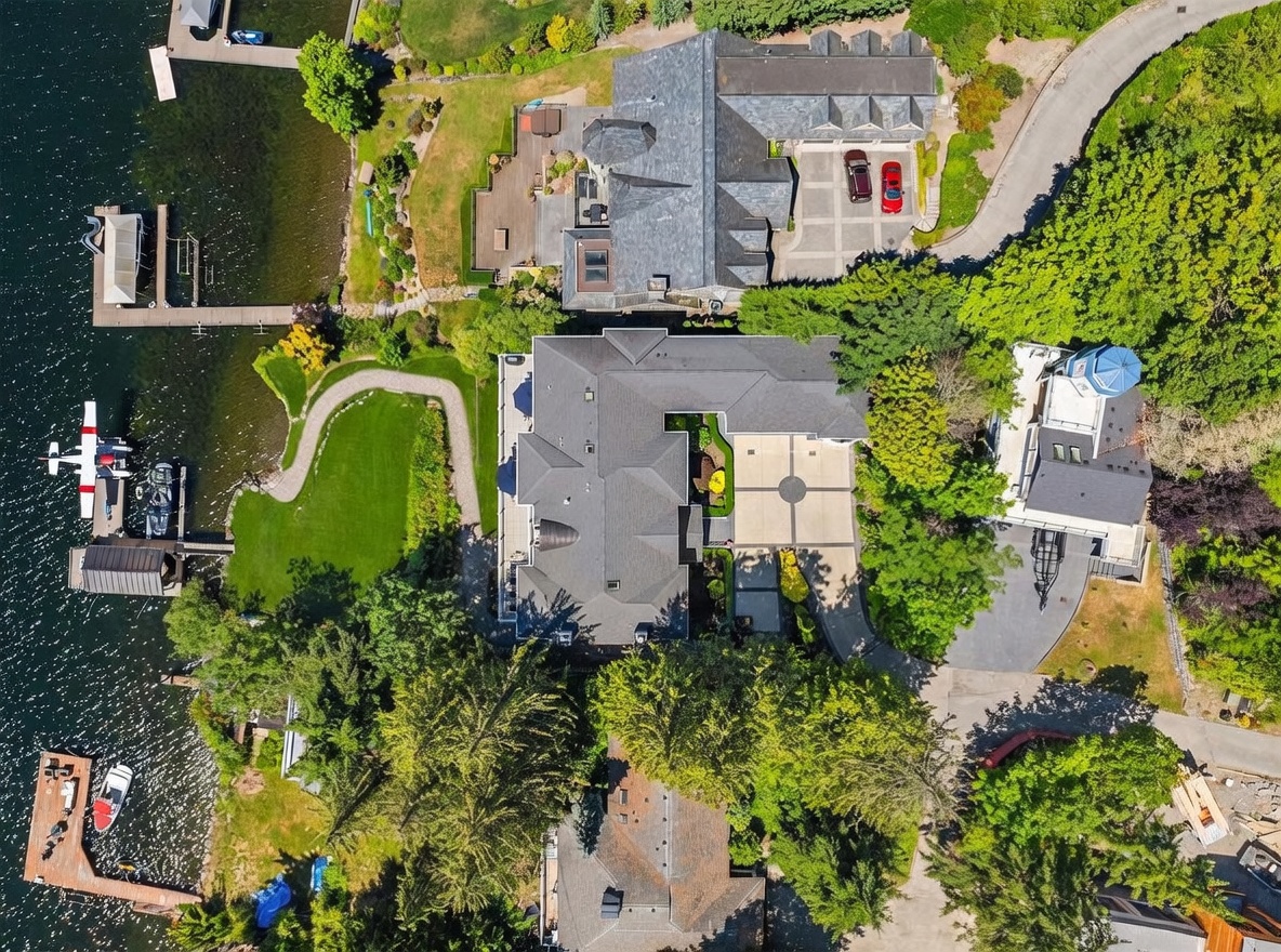 This high-altitude aerial view captures a luxurious waterfront estate featuring multiple large homes with complex rooflines, expansive paved driveways, and lush green landscaping. The property includes private docks extending into the lake, with a seaplane moored at one of the piers, emphasizing a high-end, recreational lifestyle. The perspective provides a comprehensive overview of the property's layout, proximity to the water, and surrounding mature trees.