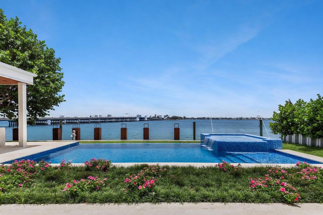 This stunning waterfront backyard features a modern infinity-edge pool and integrated spa with vibrant blue mosaic tiling. A lush garden bed filled with pink flowers and greenery borders the pool deck, while a long bridge spans the calm bay in the background. The scene captures a serene, luxurious outdoor living space perfect for relaxation and entertaining.