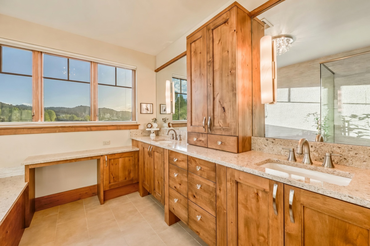 This spacious primary bathroom features a double vanity with light wood cabinetry and a light-toned stone countertop. A large mirror reflects the room, while a dedicated vanity seating area is positioned under a window that provides natural light and a view of the outdoors. The space is finished with a textured rug and elegant lighting fixtures, creating a warm and functional atmosphere.