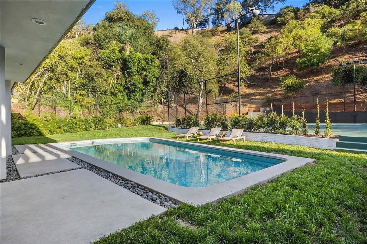 This outdoor space features a rectangular swimming pool set within a lush green lawn, bordered by a concrete patio and a decorative pebble strip. Three lounge chairs are positioned on the grass, facing a private tennis court enclosed by high fencing against a backdrop of hillside trees. The perspective captures a serene, resort-like backyard setting, emphasizing the integration of recreational amenities with natural landscaping.