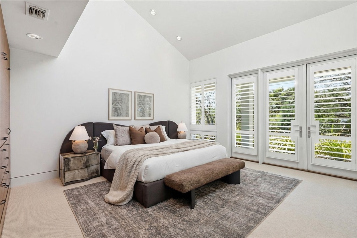 This spacious primary bedroom features a vaulted ceiling and a neutral color palette, creating an airy and serene atmosphere. A large bed with a curved, upholstered headboard serves as the focal point, complemented by a textured area rug and a bench at the foot of the bed. Tall glass doors with white shutters provide access to the outdoors, allowing natural light to fill the room.
