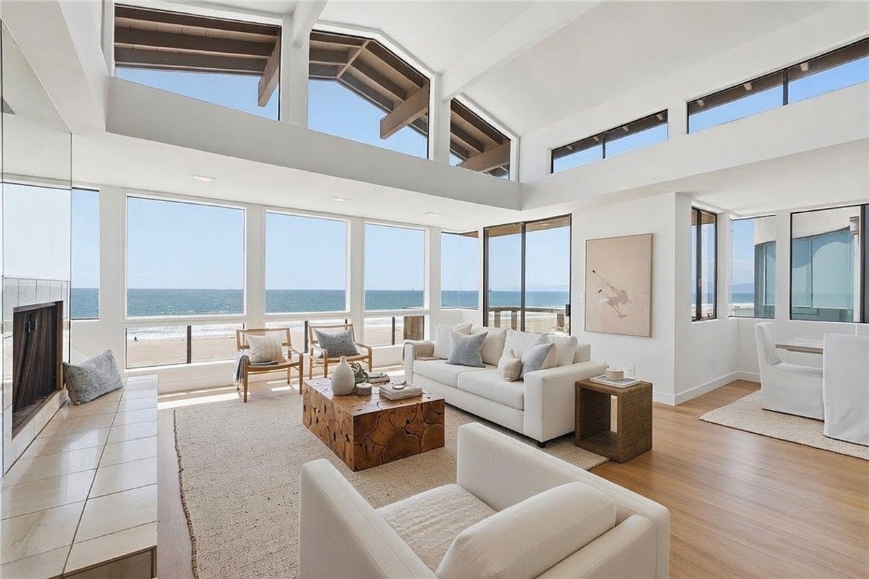 This bright and airy living room features a neutral color palette and expansive windows that provide a direct, unobstructed view of the ocean and beach. The space is furnished with a plush white sofa, two textured armchairs, and a unique, organic-shaped wooden coffee table centered on a large woven rug. The room is characterized by its high, vaulted ceilings with exposed beams and a minimalist aesthetic that emphasizes the natural light and coastal scenery.