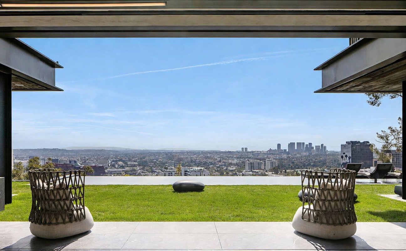 This image captures a stunning, open-air patio space that seamlessly transitions into a lush green lawn overlooking a sprawling city skyline. Two modern, woven-back lounge chairs sit on a sleek tiled floor, framing the panoramic view of the urban landscape under a clear blue sky. The architectural design emphasizes an indoor-outdoor lifestyle, creating a sophisticated and serene atmosphere perfect for luxury living.