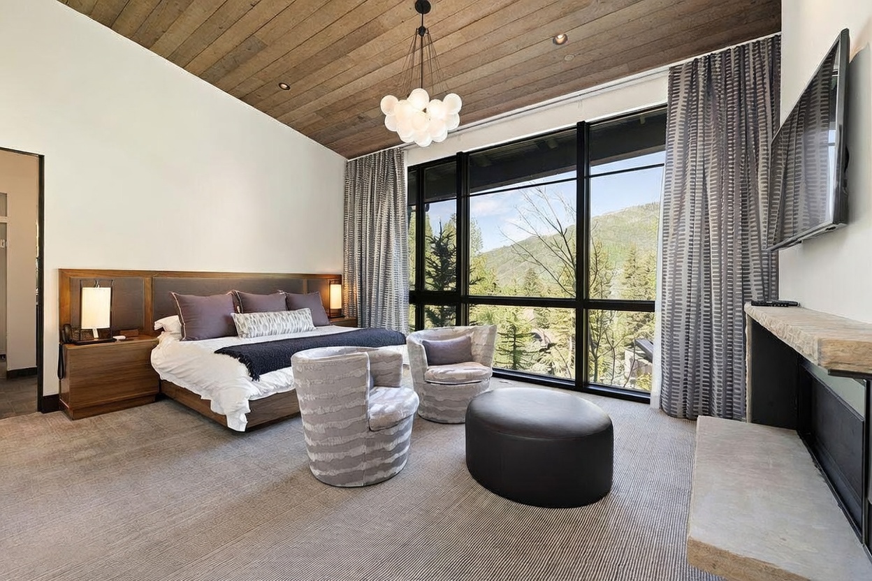 This primary bedroom features a vaulted wood-paneled ceiling and large windows that frame a scenic mountain view. The room is furnished with a king-sized bed, two patterned armchairs, and a circular ottoman, all set against a neutral color palette. A stone-clad fireplace mantel is visible on the right, adding a cozy element to the sophisticated, modern mountain aesthetic.