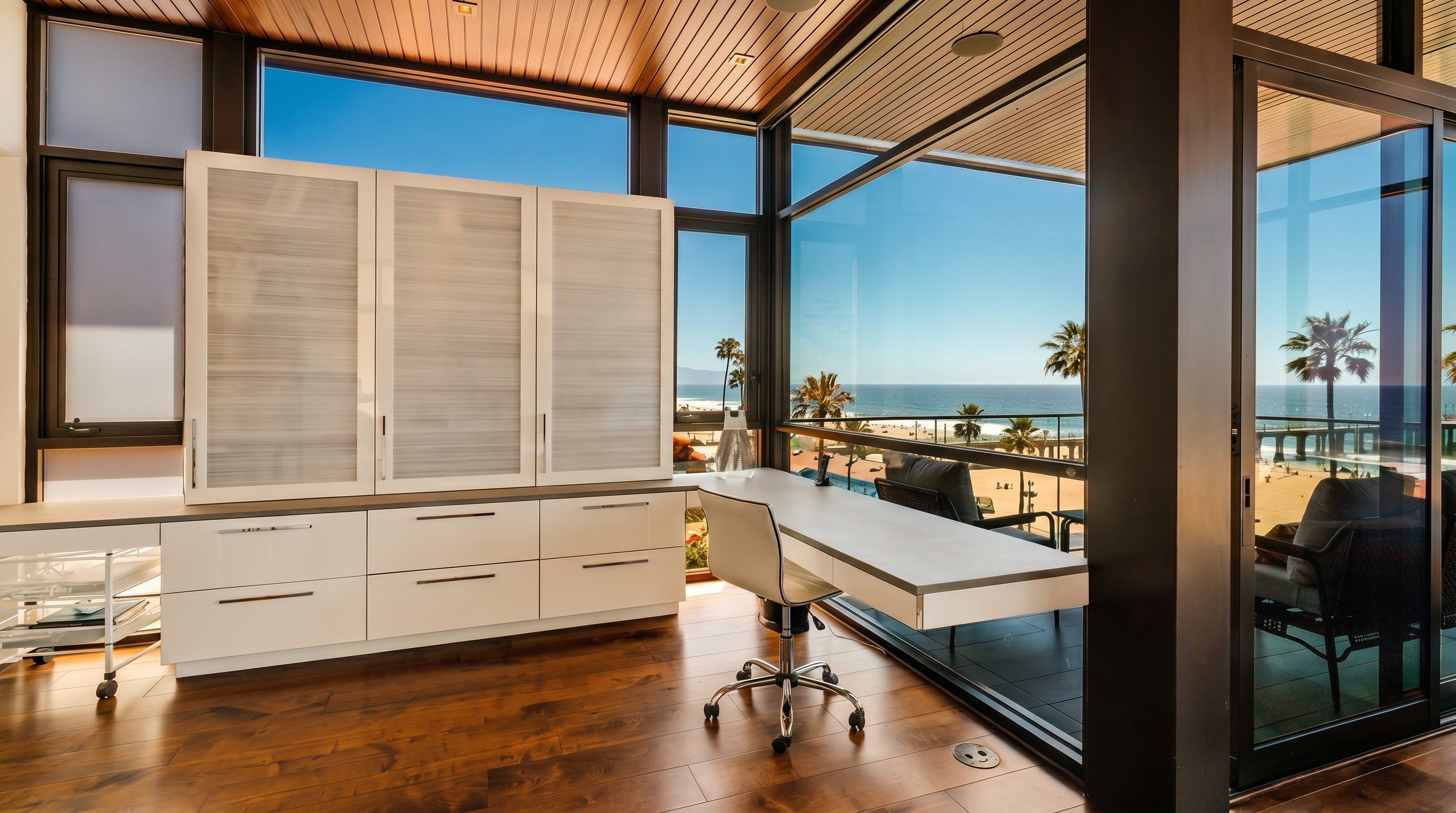 This modern home office features a sleek, minimalist desk and built-in cabinetry, set against a backdrop of expansive glass walls that offer panoramic ocean and beach views. The room is finished with warm-toned wood flooring and a matching wood-paneled ceiling, creating a sophisticated and serene workspace. The perspective captures the seamless transition between the indoor office environment and the outdoor balcony, emphasizing the property's prime coastal location.