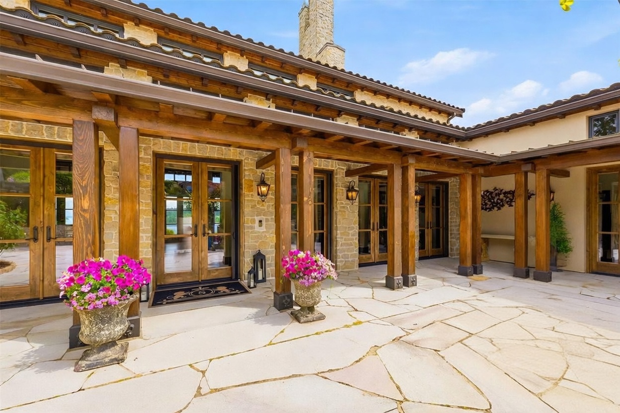 This elegant stone-paved patio features a central tiered fountain and is framed by a covered walkway with heavy timber posts and beams. The exterior walls are constructed of textured stone, complemented by multiple sets of glass-paned double doors that provide seamless indoor-outdoor access. Potted plants with vibrant pink flowers add a touch of color to the sophisticated, rustic-inspired outdoor living space.