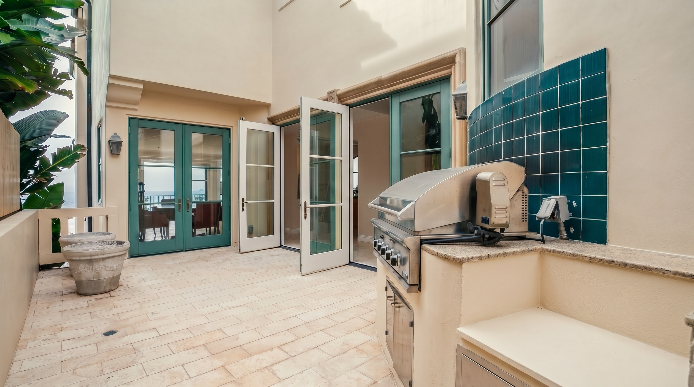 This outdoor patio space features light-colored stone pavers and a built-in stainless steel grill station with a teal tiled backsplash. Multiple sets of glass-paned French doors provide seamless access to the interior, while a large potted plant adds a touch of greenery to the corner. The perspective is from the patio looking toward the house, highlighting a functional and inviting space for outdoor dining and entertaining.