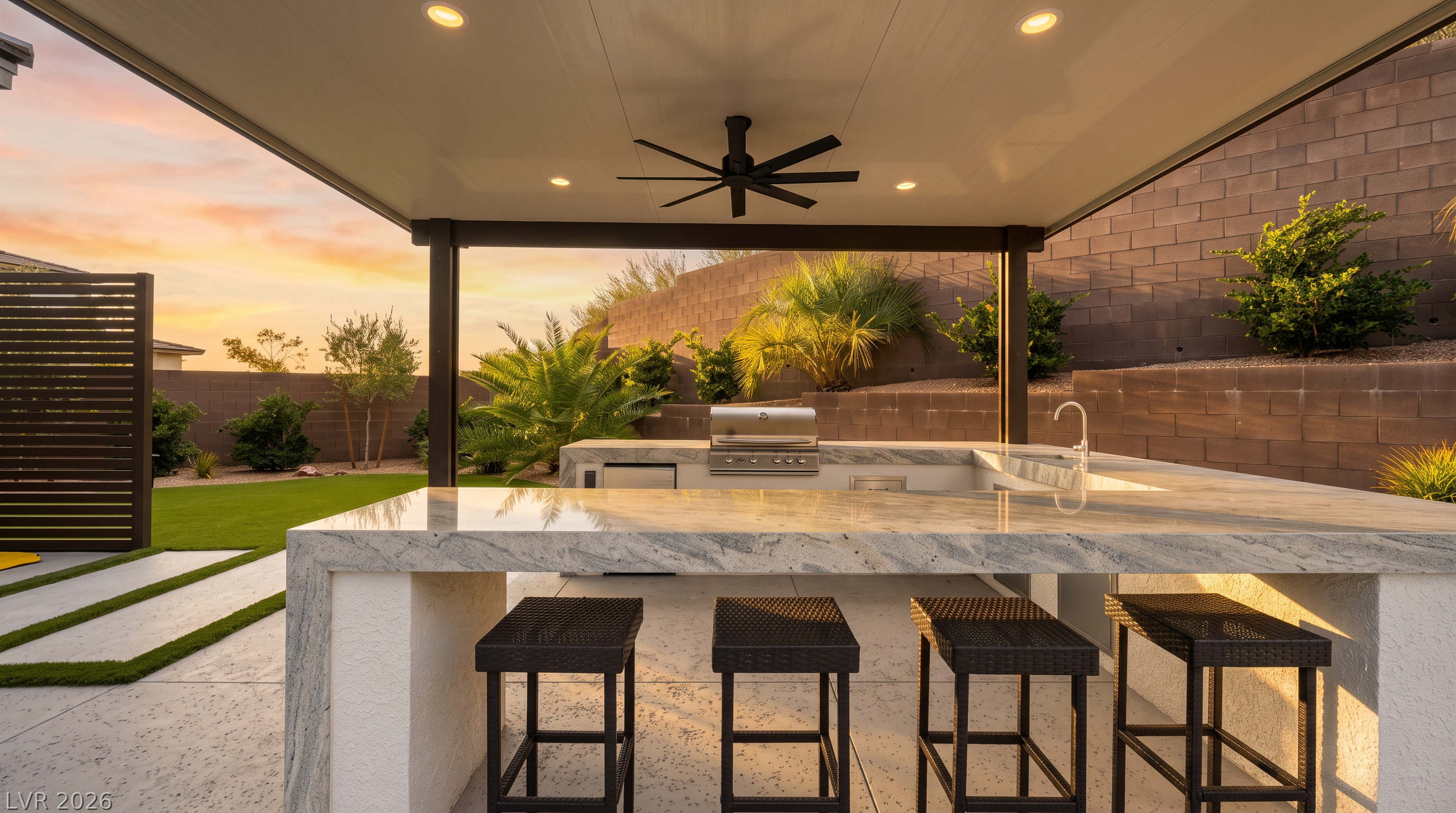 This outdoor living space features a covered patio area equipped with a built-in kitchen, including a grill, sink, and a stone-topped island with four bar stools. The area is sheltered by a modern roof structure with recessed lighting and a ceiling fan, overlooking a landscaped backyard with a lawn and tiered retaining wall. The perspective is from the patio looking outward, showcasing a clean, contemporary design ideal for outdoor entertaining.