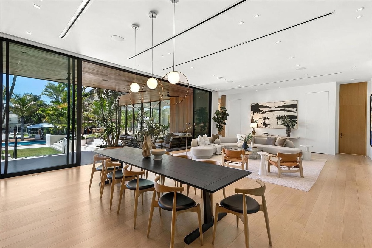 This expansive open-concept living and dining area features light wood flooring and a seamless transition to an outdoor patio through large sliding glass doors. The space is anchored by a long, dark dining table in the foreground and a comfortable seating area with a curved sofa and neutral-toned furniture in the background. Modern pendant lights hang above the dining table, while the room is bathed in natural light, creating an airy and sophisticated atmosphere.