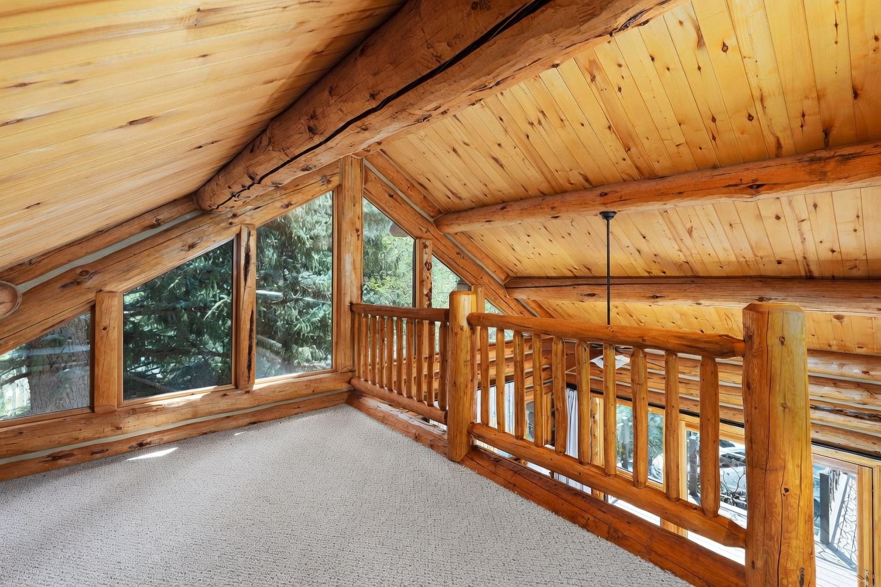This rustic loft space features a vaulted ceiling with exposed log beams and light wood paneling. A wooden railing overlooks the lower level, while large, angled windows provide a view of the surrounding trees. The area is carpeted, creating a cozy and elevated retreat within the home.